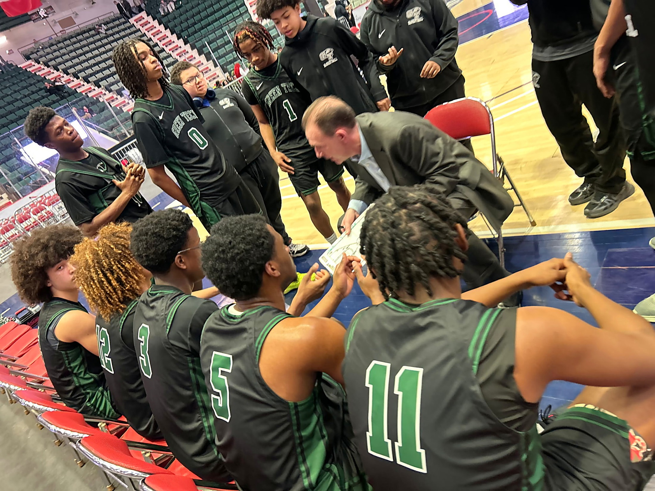 Green Tech Eagles basketball players gathered around their head coach during a timeout huddle as he draws up a play
